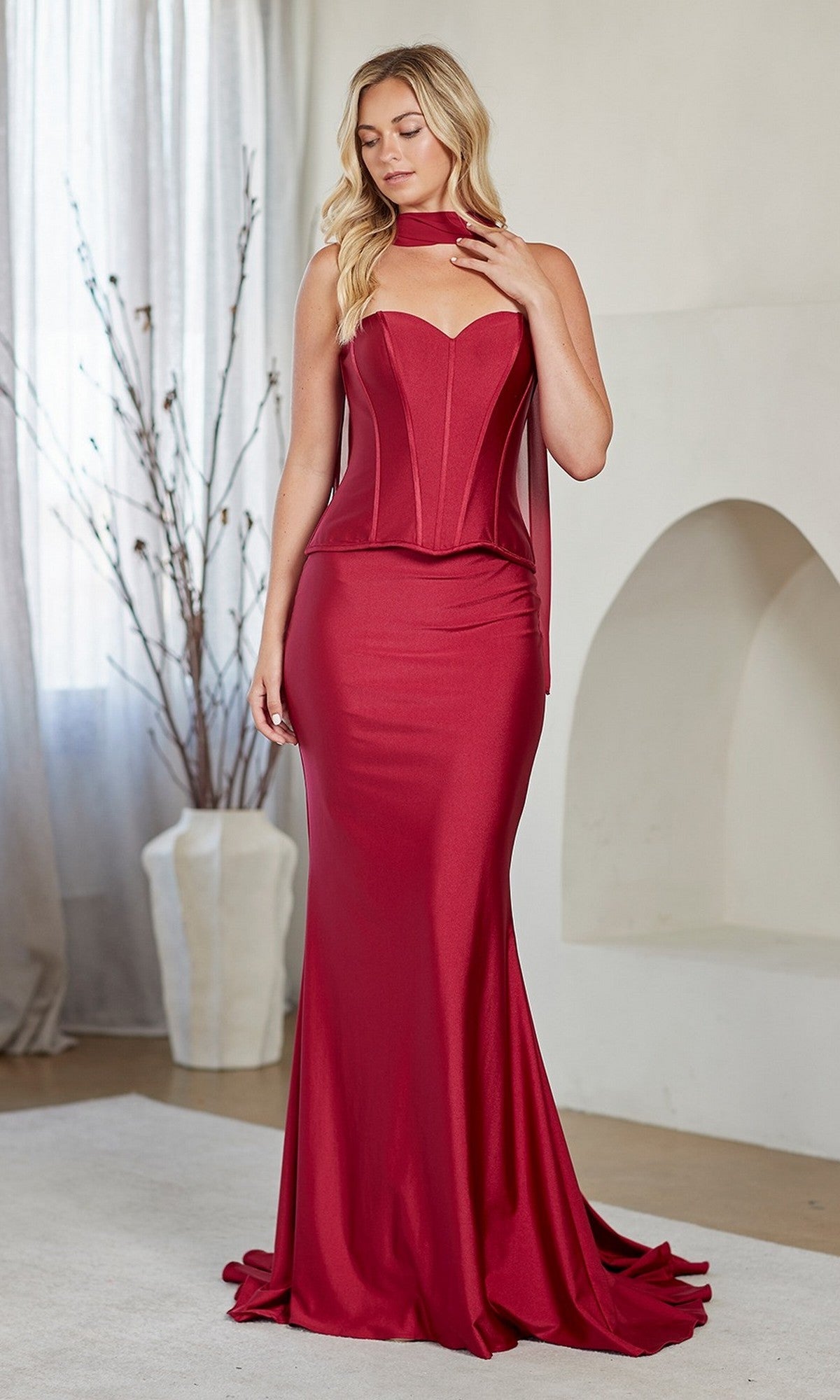 A woman stands indoors wearing the Amelia Couture JQ1105 Long Prom Dress, a fitted, strapless red satin gown with a sweetheart neckline, floor-length skirt, and a matching neck scarf.